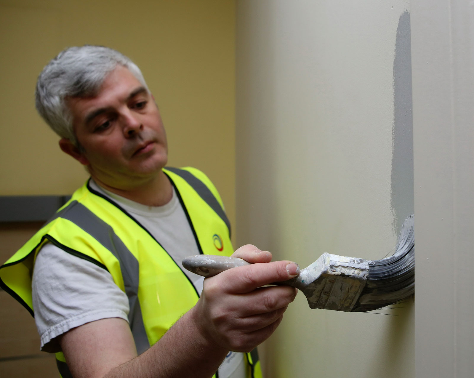 Photograph of a painter wearing hi-viz jacket and applying paint with a brush to a wall.